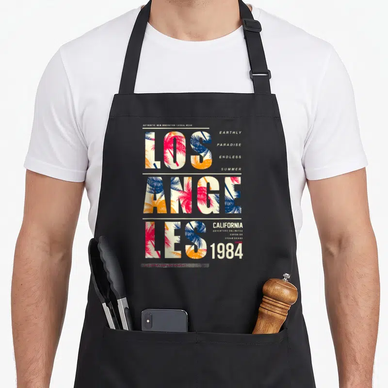 Tablier de cuisine noir avec typographie Los Angeles remplie de palmes roses et bleues ainsi que les mots California 1984.