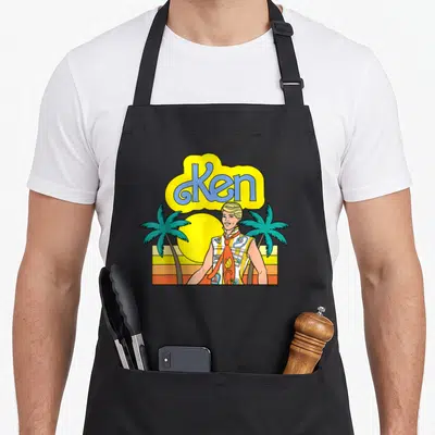 Tablier de cuisine noir avec illustration colorée de Ken, palmiers et soleil couchant aux teintes chaudes.