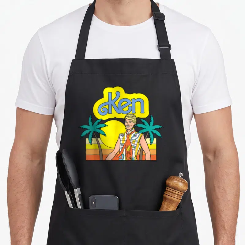 Tablier de cuisine noir avec illustration colorée de Ken, palmiers et soleil couchant aux teintes chaudes.