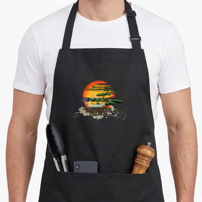 Tablier de cuisine noir avec illustration d'un arbre bonsaï vert devant un soleil orange et des motifs floraux blancs.