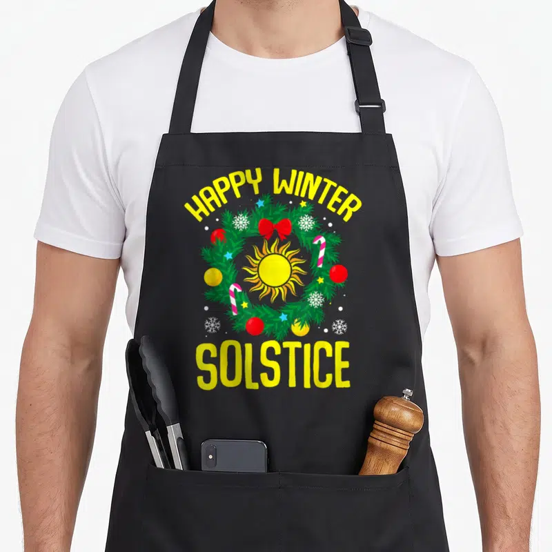 Tablier de cuisine noir avec couronne de sapin, soleil jaune et texte Happy Winter Solstice en lettres jaunes.