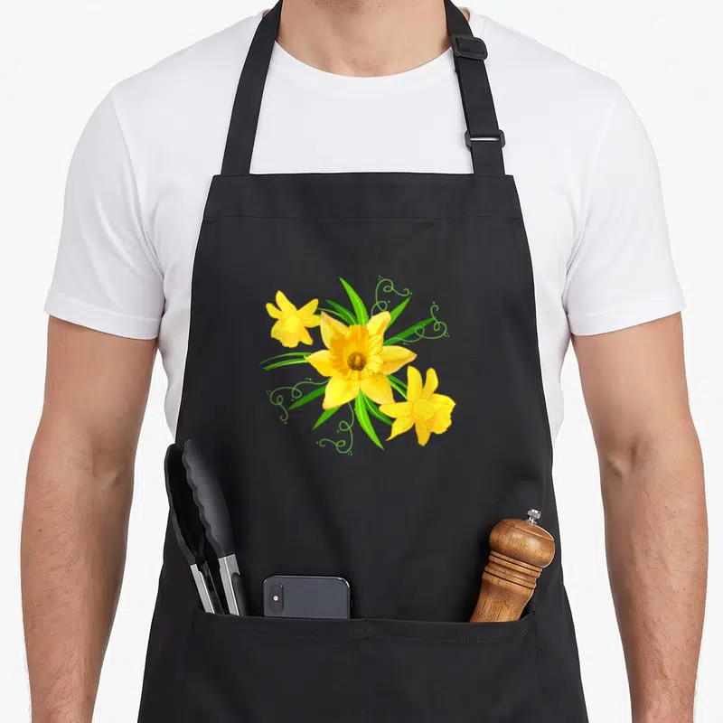 Tablier de cuisine noir avec un bouquet de jonquilles jaunes, de feuilles vertes et de volutes décoratives.