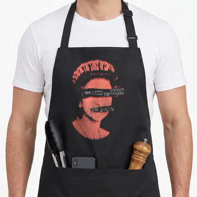 Tablier de cuisine noir avec portrait stylisé en timbre rouge et texte God Save the Queen barré sur les yeux.