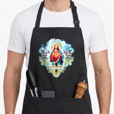 Tablier de cuisine noir avec une illustration colorée de Jésus-Christ, un cœur rayonnant, une croix dorée et des arabesques végétales.
