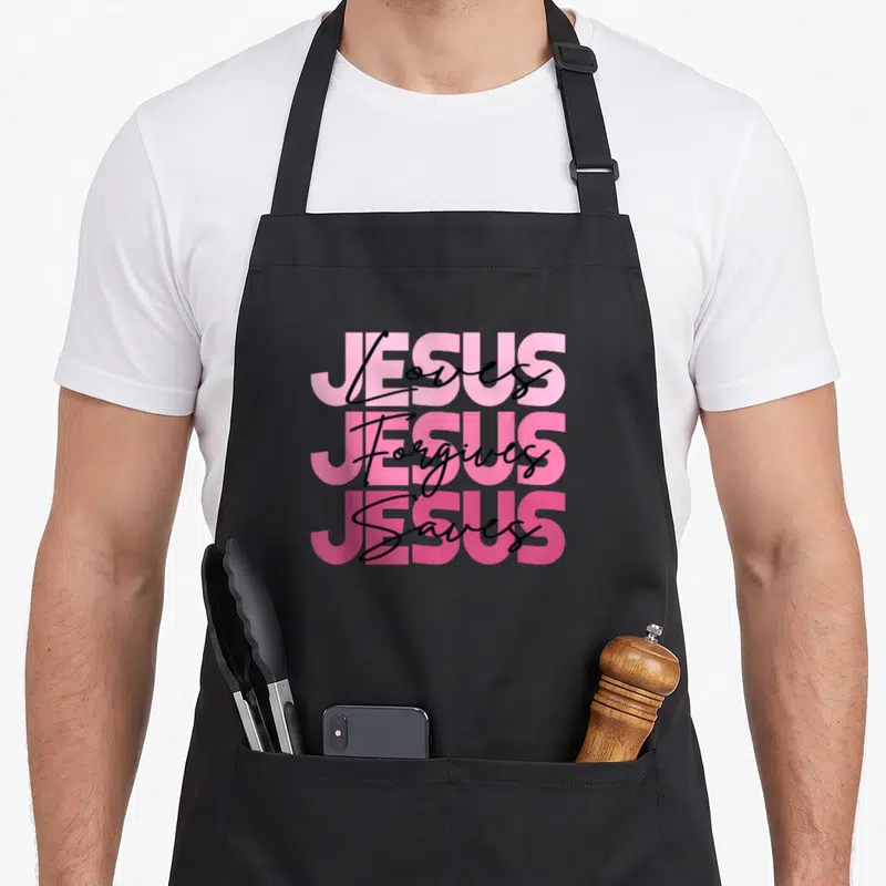 Tablier de cuisine noir avec l'inscription Jésus aime pardonne sauve en typographie rose et noire superposée.