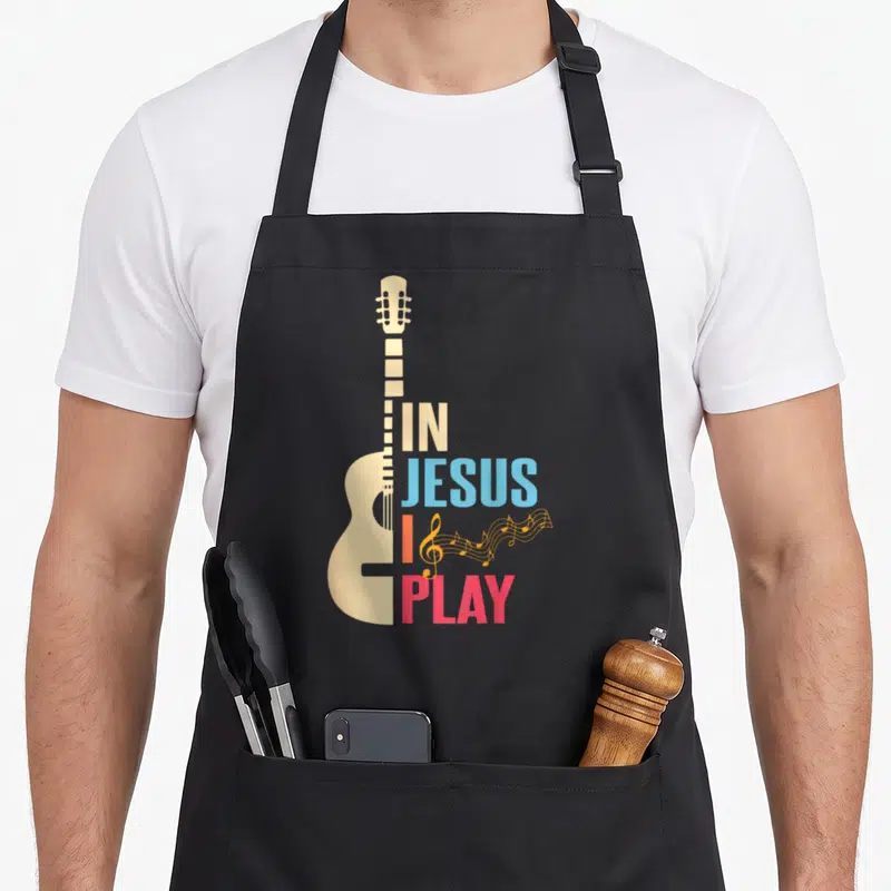Tablier de cuisine noir avec illustration de guitare beige et texte multicolore En Jésus je joue.