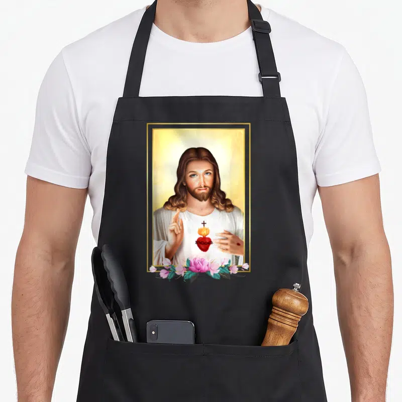 Tablier de cuisine noir avec une illustration du Sacré-Cœur de Jésus entouré de roses roses et d'un cadre doré.