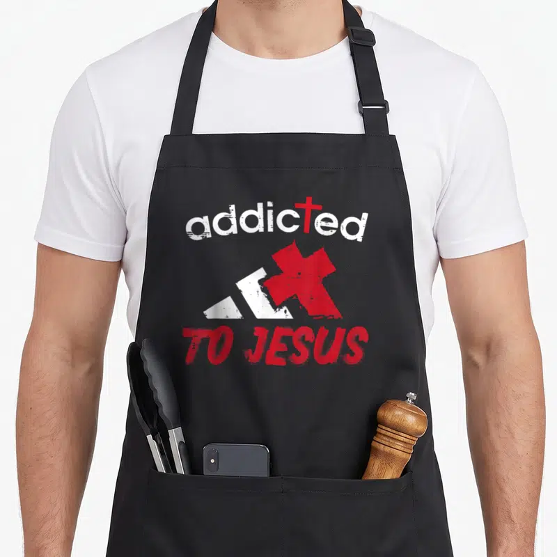 Tablier de cuisine noir avec l'inscription Addicted To Jesus en rouge et blanc accompagnée d'une croix et d'un logo graphique stylisé.