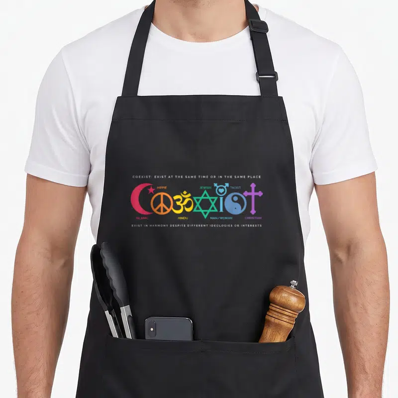 Tablier de cuisine noir avec les mots Coexist et des symboles religieux colorés sur le devant.