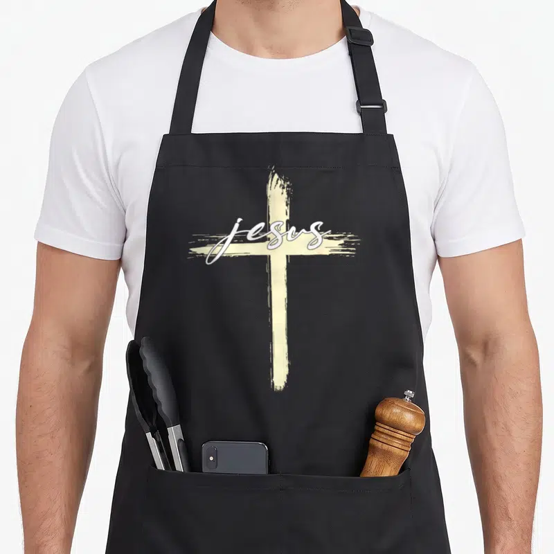 Tablier de cuisine noir avec une croix beige et le mot Jesus écrit en blanc au centre.