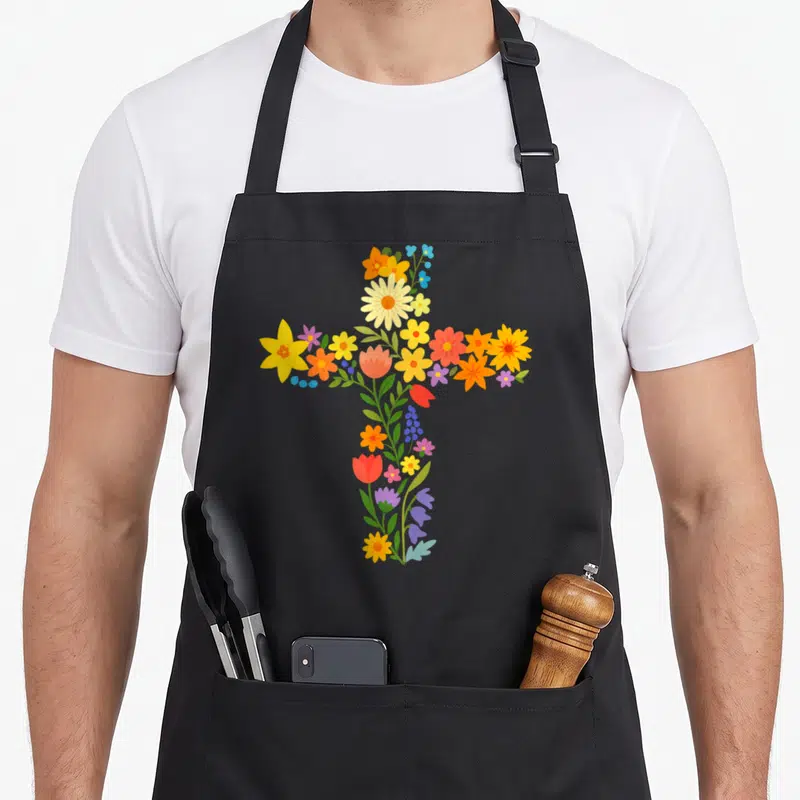 Tablier de cuisine noir avec une croix composée de fleurs multicolores et de feuilles vertes stylisées.