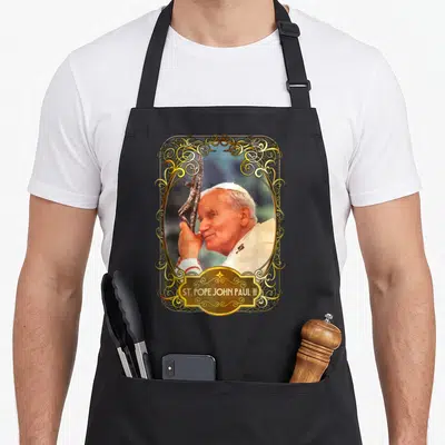 Tablier de cuisine noir avec portrait de St. Pope John Paul II encadré de motifs dorés et une croix en bois.
