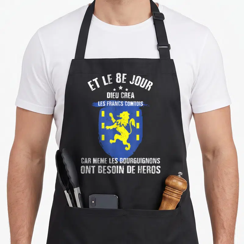 Tablier de cuisine noir avec un blason bleu et jaune représentant un lion et du texte blanc humoristique sur les Francs Comtois.
