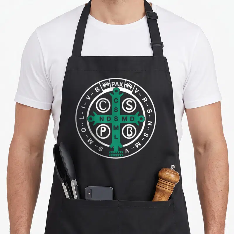 Tablier de cuisine noir avec le sceau de Saint Benoît en vert et blanc, croix et inscriptions latines.