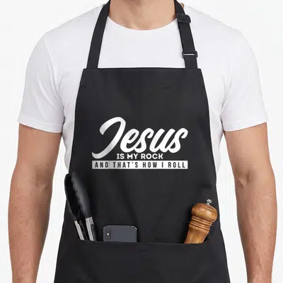 Tablier de cuisine noir avec le texte blanc « Jesus IS MY ROCK AND THAT'S HOW I ROLL » imprimé au centre du torse.