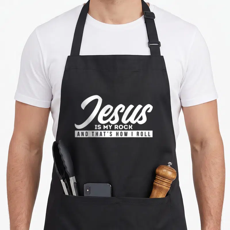 Tablier de cuisine noir avec le texte blanc « Jesus IS MY ROCK AND THAT'S HOW I ROLL » imprimé au centre du torse.
