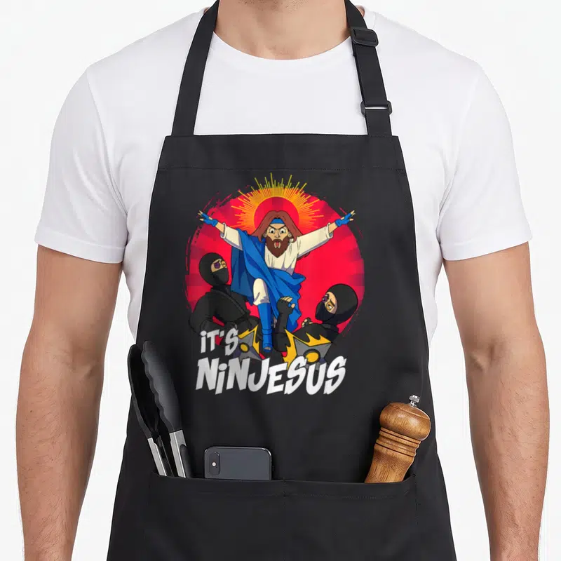 Tablier de cuisine noir avec illustration colorée d'un Jésus ninja et du texte It's Ninjesus en blanc.