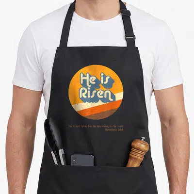 Tablier de cuisine noir avec logo circulaire orange affichant He is Risen en lettres bleues et le texte Matthew 28:6 en bas.
