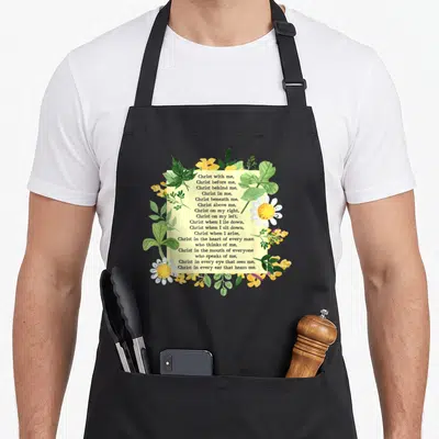 Tablier de cuisine noir avec prière chrétienne encadrée de fleurs blanches et de feuilles vertes sur fond crème.