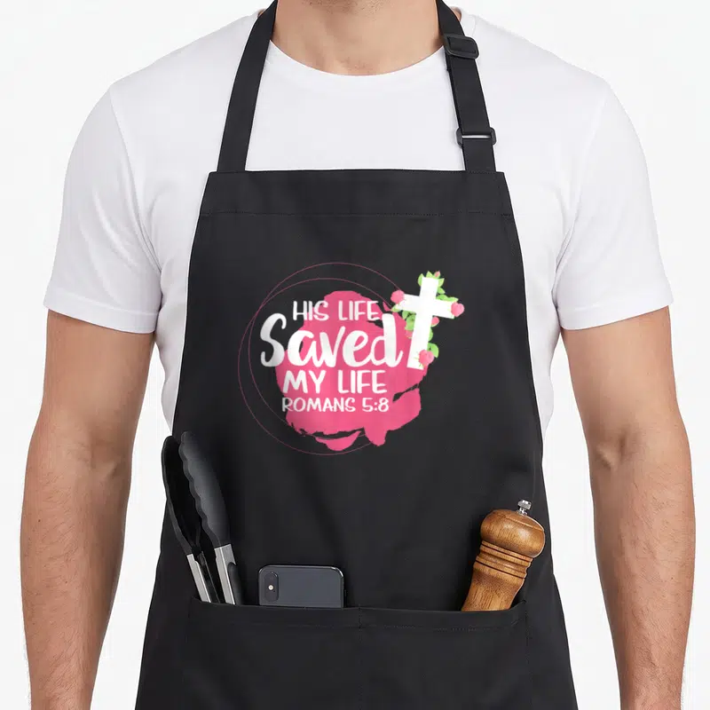 Tablier de cuisine noir avec inscription His Life Saved My Life, croix blanche et fleurs roses sur tache rose.
