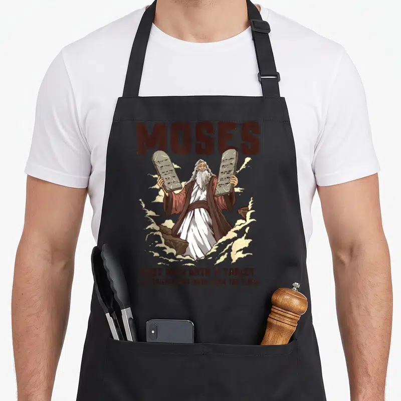Tablier de cuisine noir avec illustration de Moïse tenant deux tablettes et le texte Moses en haut du design imprimé.