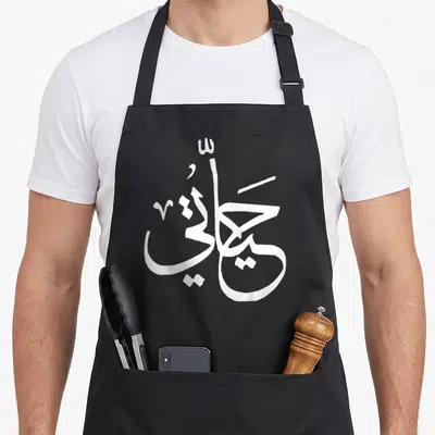 Tablier de cuisine noir avec une grande calligraphie arabe blanche stylisée au centre du vêtement.