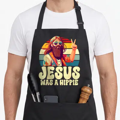 Tablier de cuisine noir avec illustration d'un Jésus hippie aux longs cheveux et texte Jesus was a hippie en dessous.