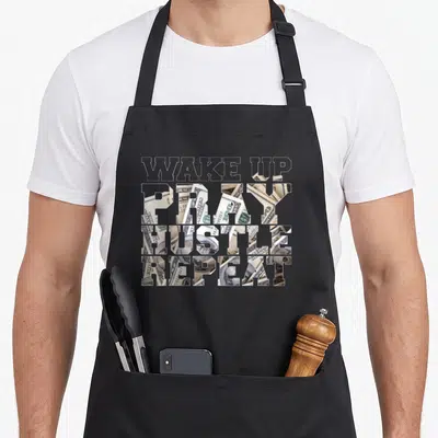 Tablier de cuisine noir avec des mots Wake Pray Hustle Repeat remplis de billets de dollars américains superposés.