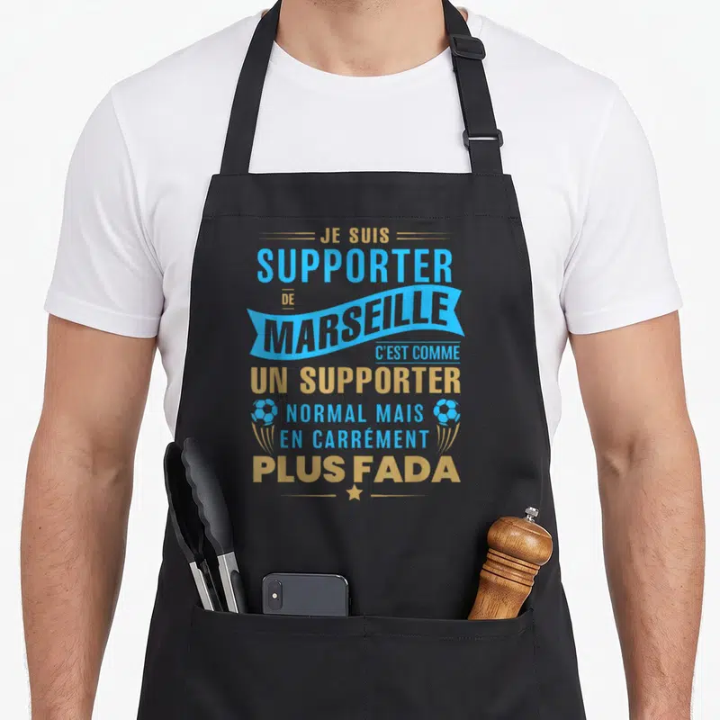 Tablier de cuisine noir avec inscription supporteur de Marseille plus fada en bleu et or, orné de ballons de foot stylisés.