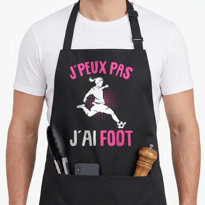 Tablier de cuisine noir avec silhouette de jeune footballeuse et texte J'peux pas j'ai foot en rose et blanc.