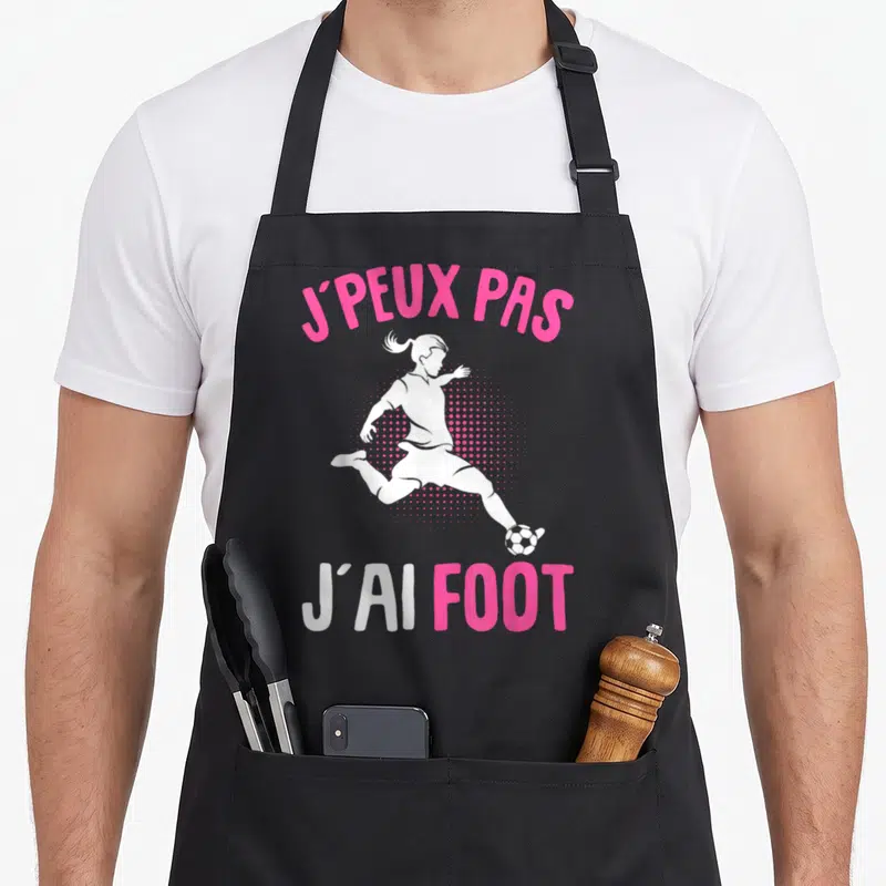 Tablier de cuisine noir avec silhouette de jeune footballeuse et texte J'peux pas j'ai foot en rose et blanc.