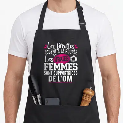 Tablier de cuisine noir avec texte blanc et rose affirmant que les vraies femmes sont supportrices de l'OM près d'un ballon de foot.