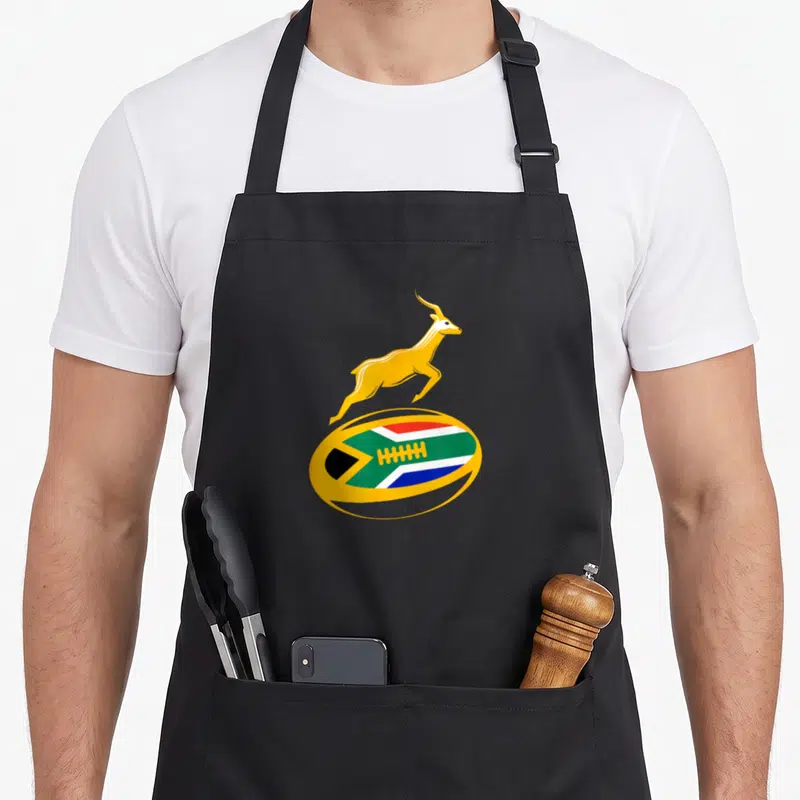 Tablier de cuisine noir avec un ballon de rugby aux couleurs du drapeau sud-africain et un springbok doré sautant par-dessus.