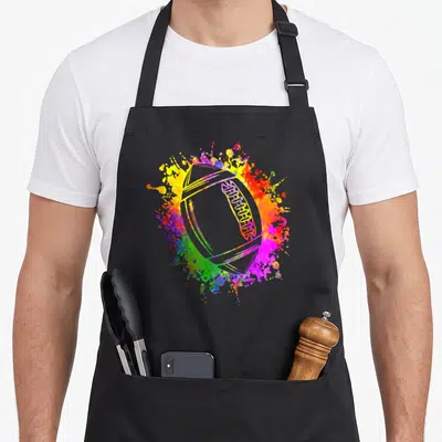 Tablier de cuisine noir avec un ballon de rugby entouré d'éclats de peinture jaune, rose et vert vif.