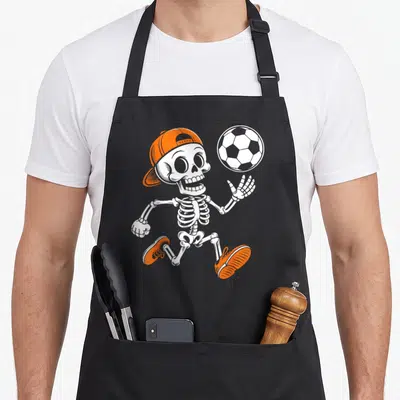 Tablier de cuisine noir avec squelette portant casquette et chaussures orange jouant au ballon blanc.