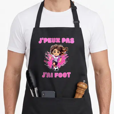 Tablier de cuisine noir avec une jeune joueuse de foot et le texte J'peux pas j'ai foot en lettres roses.