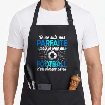 Tablier de cuisine noir avec inscription humoristique sur le football féminin et un ballon graphique central.