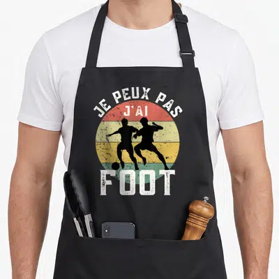 Tablier de cuisine noir avec silhouette de joueurs de foot et texte Je peux pas j'ai foot sur fond de soleil coloré.