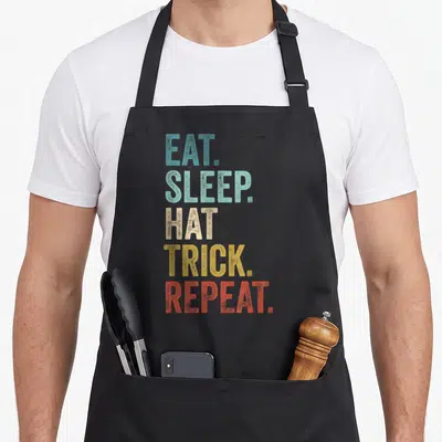 Tablier de cuisine noir avec le texte Eat Sleep Hat Trick Repeat en lettres multicolores verticales.