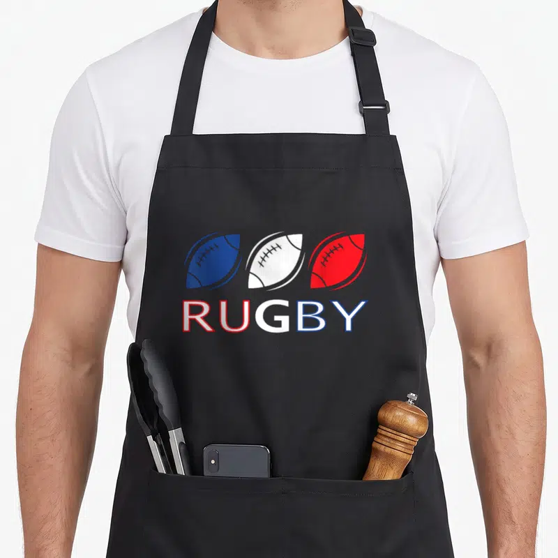 Tablier de cuisine noir avec trois ballons de rugby bleus blancs rouges et le mot RUGBY imprimé en bas.