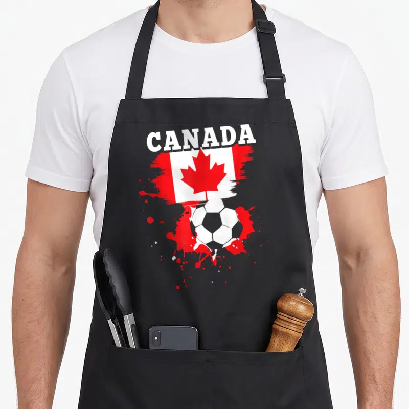 Tablier de cuisine noir avec inscription CANADA, drapeau canadien et ballon de football entouré de taches rouges.