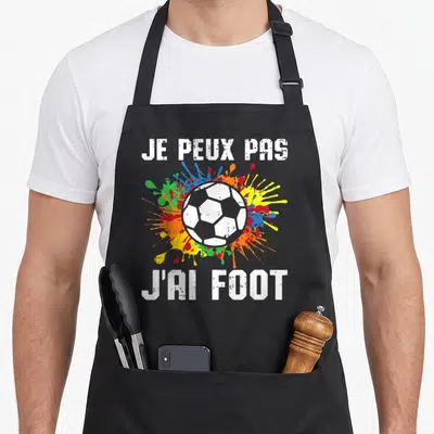 Tablier de cuisine noir avec un ballon de foot blanc et des éclaboussures multicolores derrière le texte blanc.