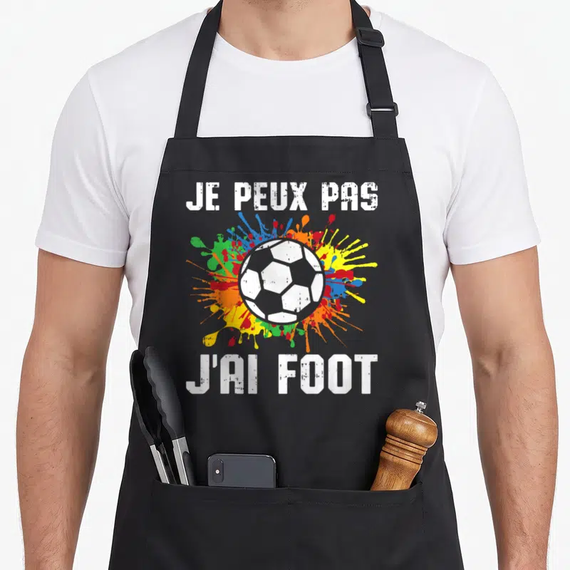 Tablier de cuisine noir avec un ballon de foot blanc et des éclaboussures multicolores derrière le texte blanc.