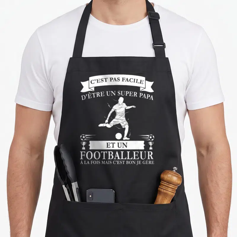 Tablier de cuisine noir avec silhouette de footballeur et texte blanc C'est pas facile d'être un super papa et un footballeur.