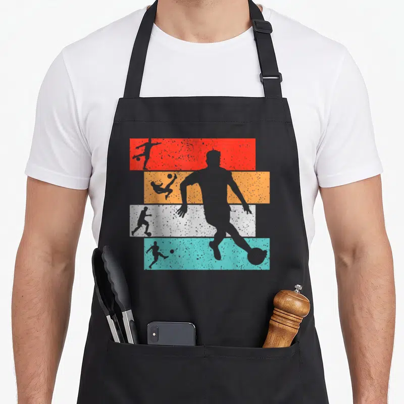 Tablier de cuisine noir avec silhouettes de footballeurs sur bandes rouges, oranges, blanches et bleues texturées.