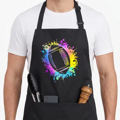 Tablier de cuisine noir avec un ballon de rugby entouré d'éclaboussures de peinture rose, jaune et bleue.