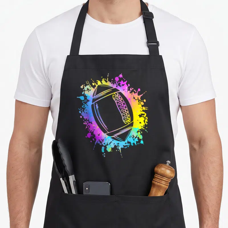 Tablier de cuisine noir avec un ballon de rugby entouré d'éclaboussures de peinture rose, jaune et bleue.
