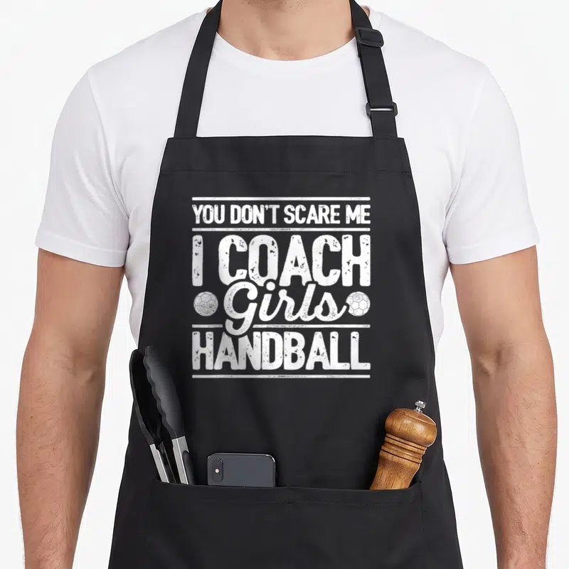 Tablier de cuisine noir avec inscription blanche I Coach Girls Handball et deux ballons de handball stylisés.
