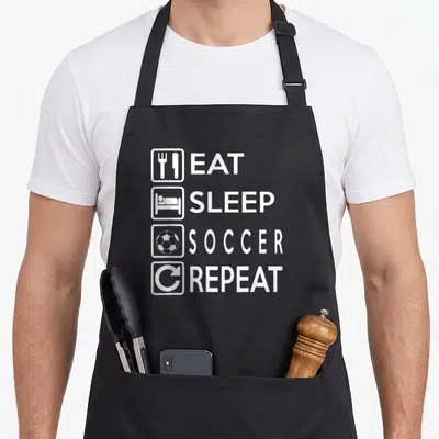 Tablier de cuisine noir avec texte blanc Eat Sleep Soccer Repeat et icônes associées.