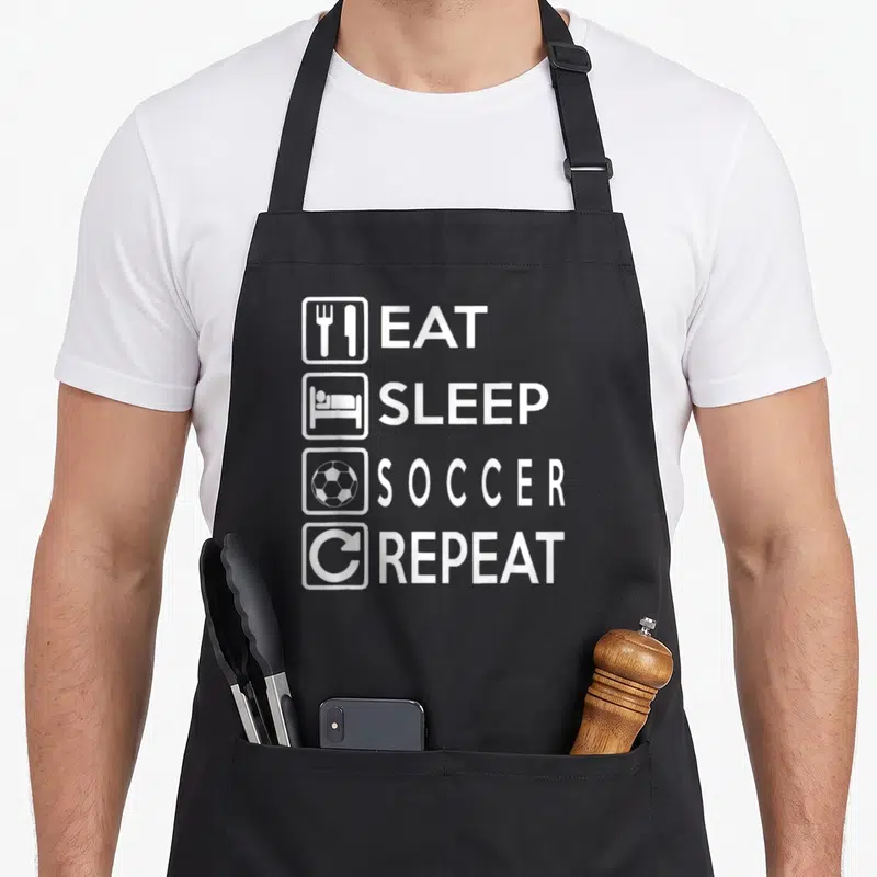 Tablier de cuisine noir avec texte blanc Eat Sleep Soccer Repeat et icônes associées.